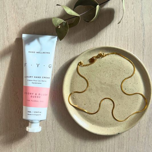 Slim gold snake chain necklace by Little Nell laid beside an FYG Peony & Blush Suede hand cream.