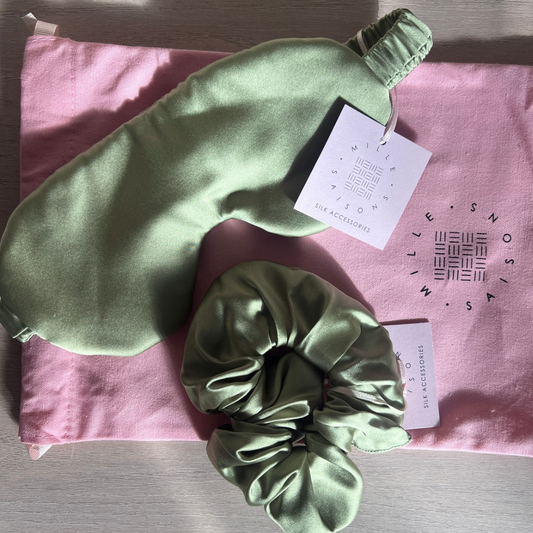A pistachio green silk sleep mask and matching scrunchie set from Mille Saisons, displayed on a pink branded fabric pouch over a light wood surface.